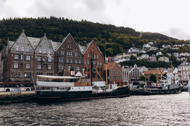 Old ships moored in front of the Bryggen wharf in Bergen, the pointed timber gables below a wooded hillside.