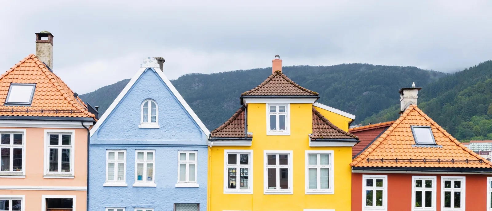 Four painted timber house gables in a row in Bergen, each a different colour and roof shape, with a forested ridge behind them.
