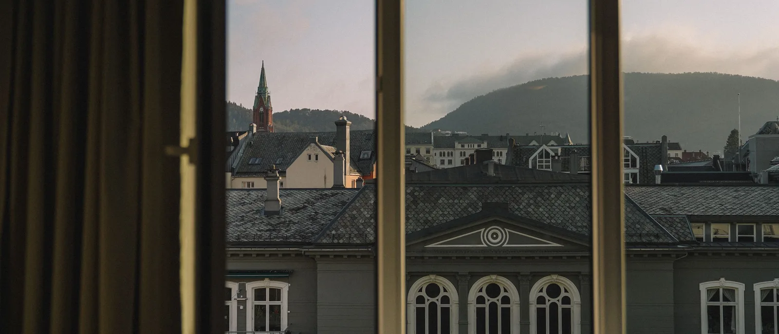 Bergen rooftops seen through the glazing bars of a window at dusk, the arched windows of a pale grey building below, a green copper church spire and a wooded ridge beyond.