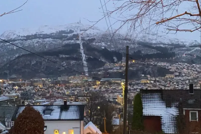 A snowy lane between garden hedges in a Bergen suburb at dusk, lit windows in a white house and the city beyond.