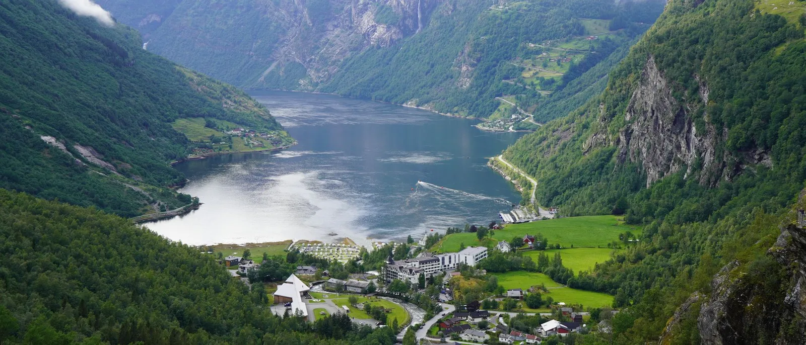 Geiranger seen from high on the valley side where the fjord ends, low cloud on the green mountain walls and a hairpin road on the far slope. By Zachary Baltimore.
