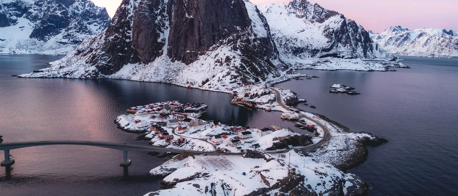 The red rorbu cabins of Hamnøy in Lofoten on their stilts, snow on the peaks behind and a bridge crossing the sound.