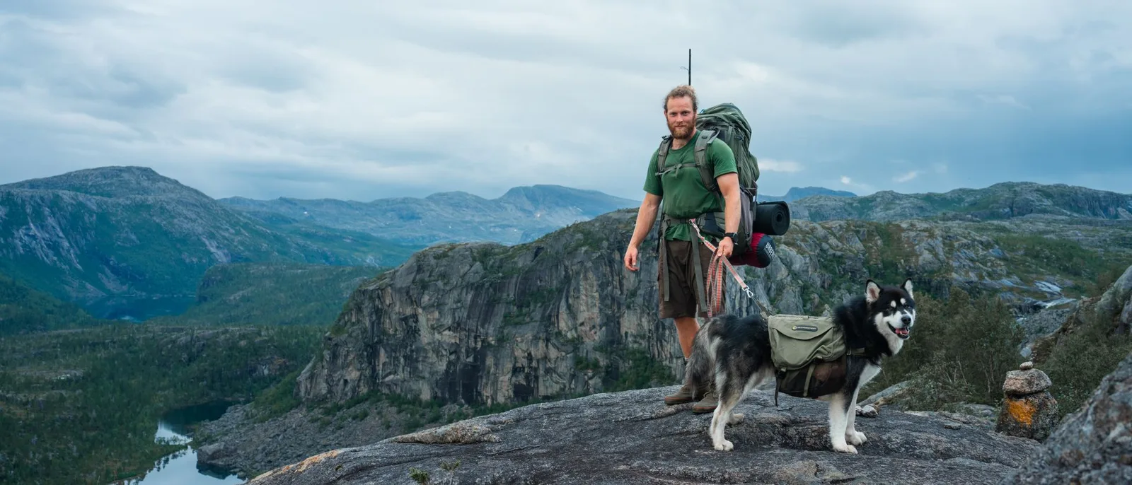 A hiker and a sled dog on a ridge in Nordland, bare mountains and a stretch of water behind them.
