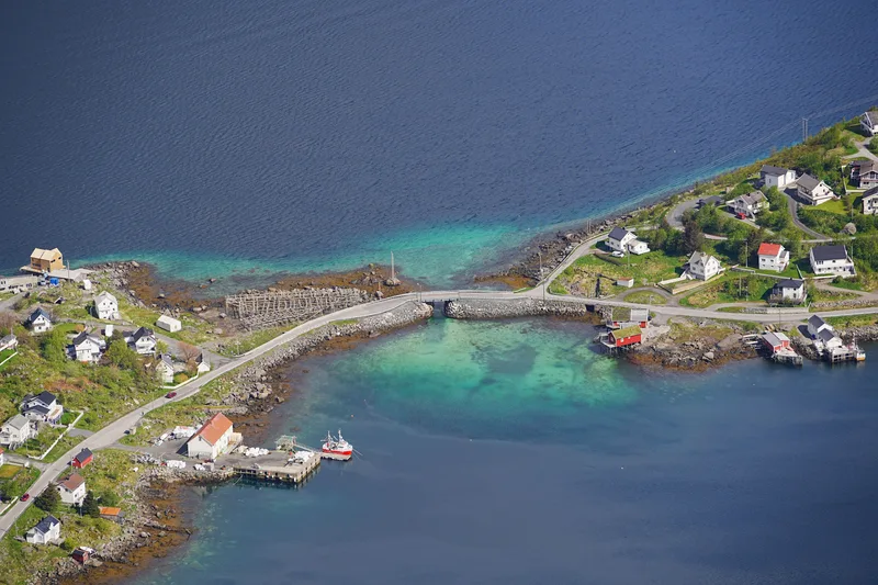 An aerial view of a Lofoten fishing village where a low stone causeway and a small bridge join two islands across shallow green water.