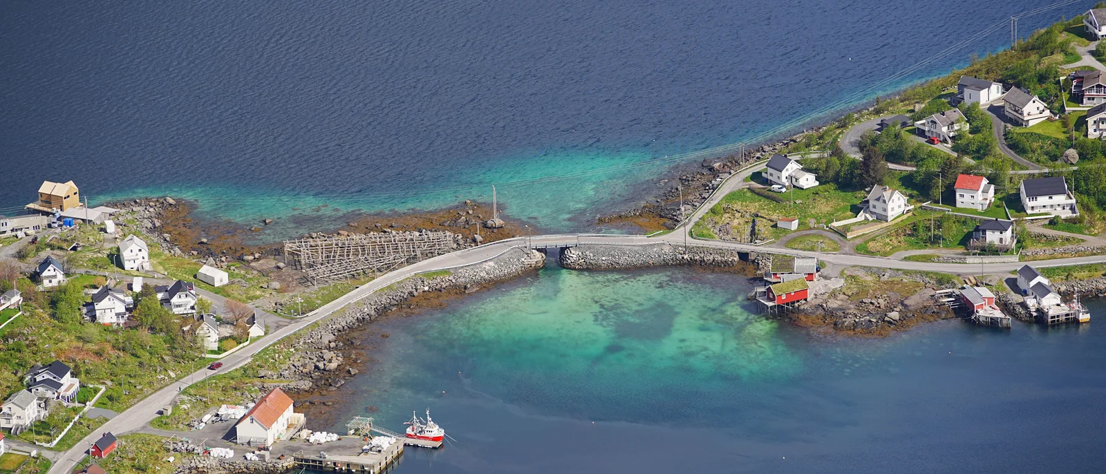 An aerial view of a Lofoten fishing village, a low causeway and a small bridge carrying one road between two islands across pale green water.