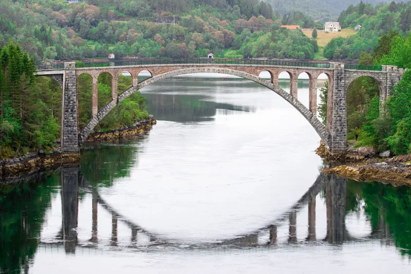 A stone arch bridge across a still fjord, its arches reflected in the water between wooded banks.