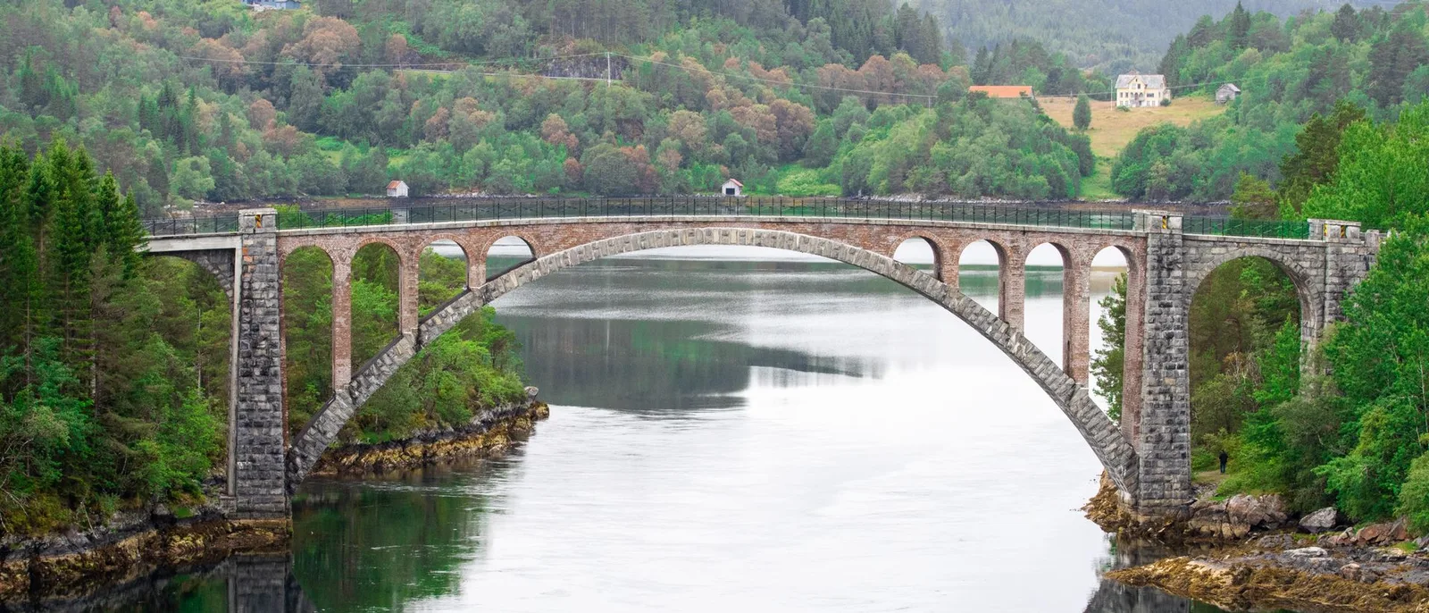 A stone arch bridge across a still fjord, its arches reflected in the water between wooded banks.