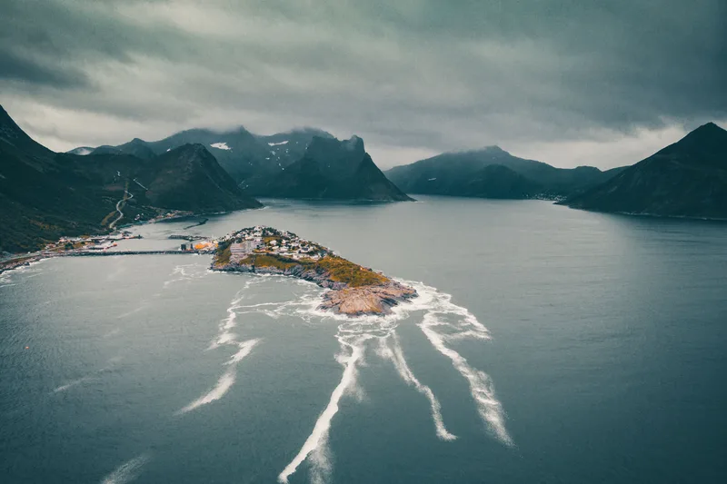 A dark aerial view of a northern Norwegian fjord under heavy cloud, a small settlement on a spit of land between steep grey mountains.