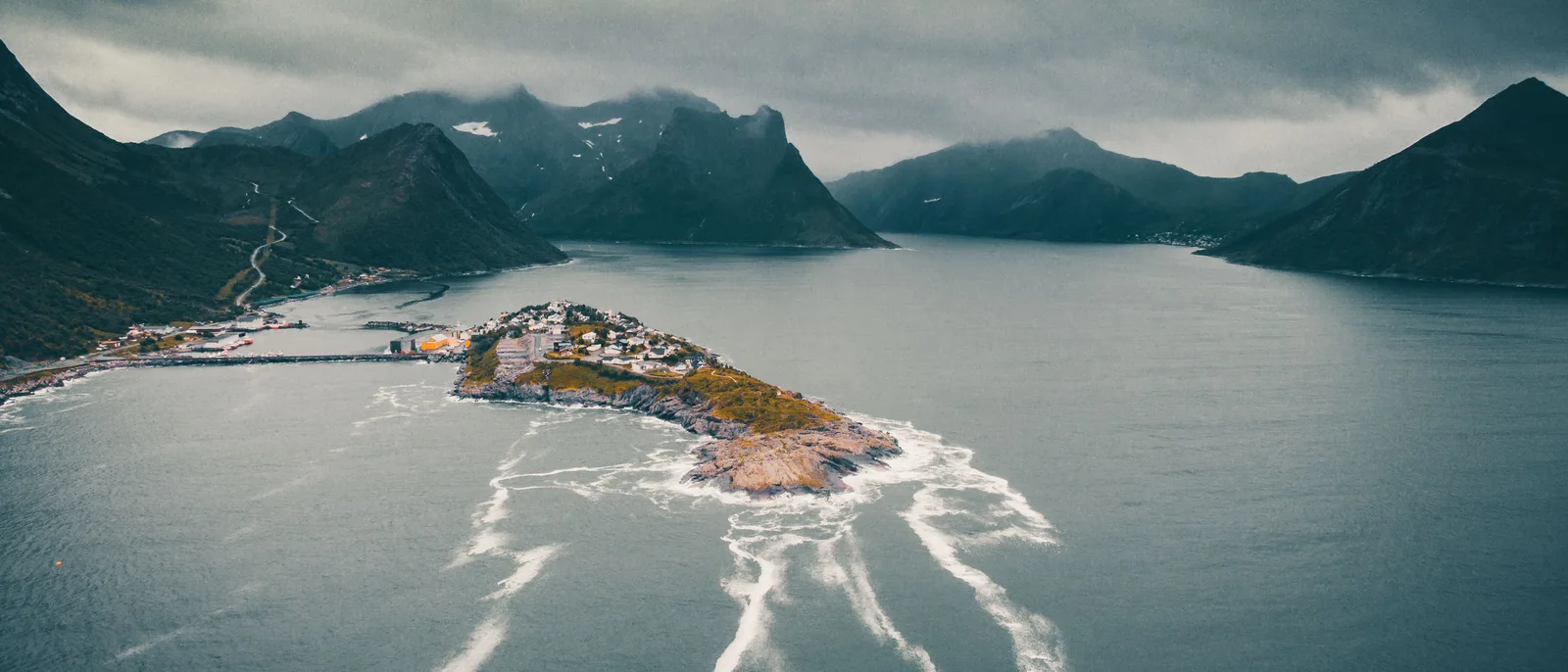 A dark aerial view of a northern Norwegian fjord under heavy grey cloud, a small settlement on a low spit of land between steep mountains, white water streaking the sea around it.
