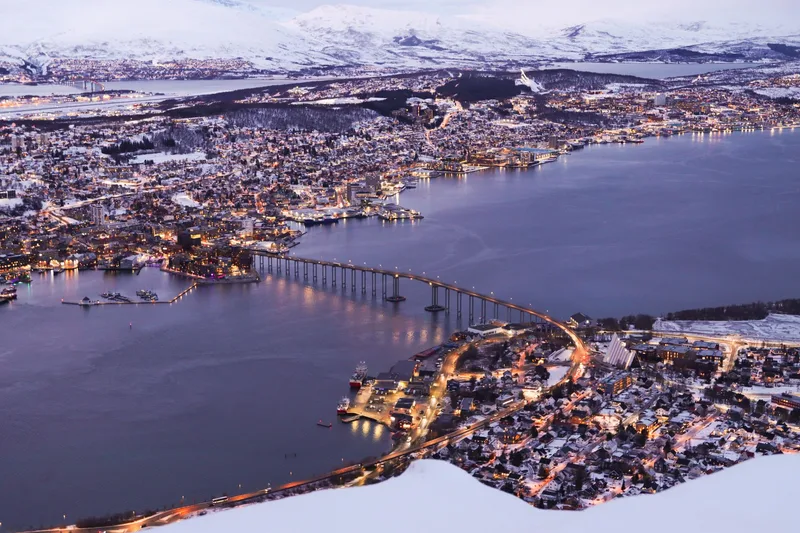 Tromsø at blue hour, the bridge lit across the sound with the lights of the town and snow on the hills behind it.