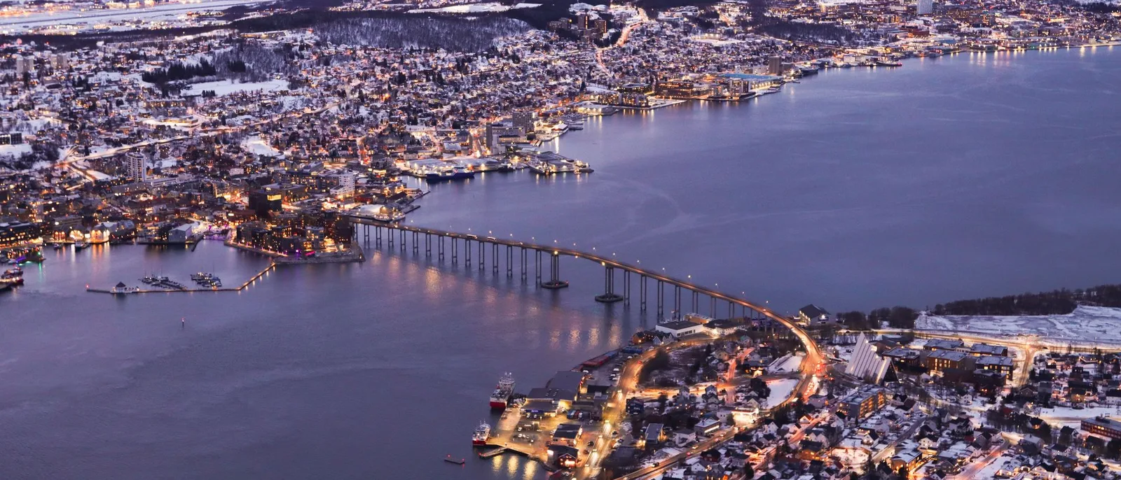 Tromsø at blue hour, the bridge lit across the sound with the lights of the town and the snow on the hills behind it.