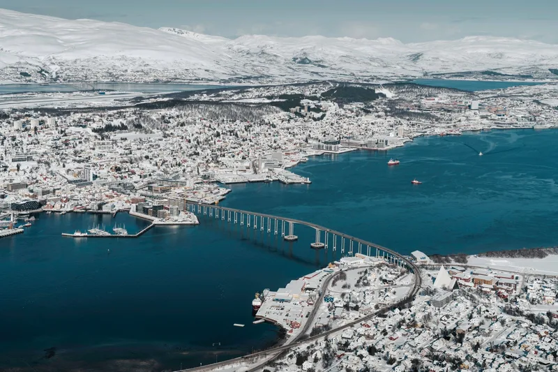 Tromsø in winter seen from above, the Tromsø Bridge crossing the blue sound between the snow-covered island and mainland.