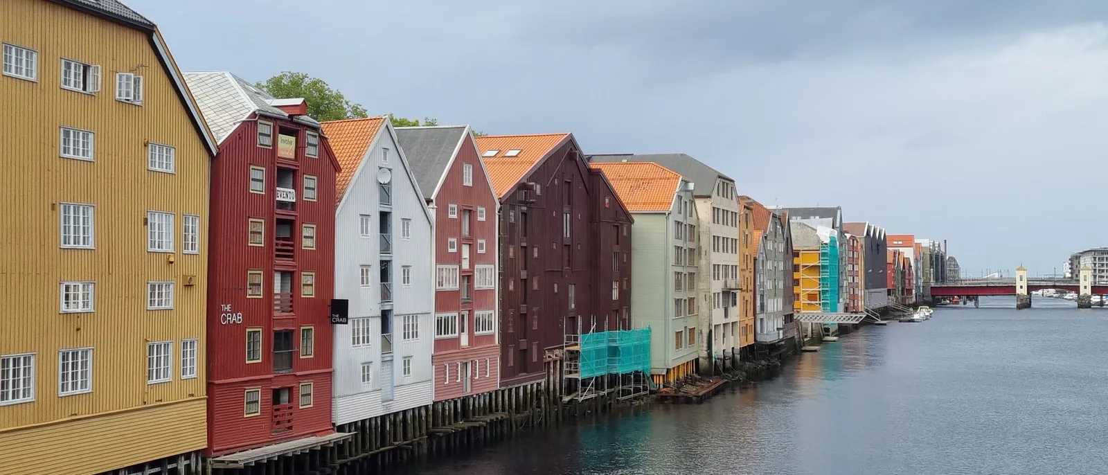Painted timber wharf houses on stilts along the Nidelva river in Trondheim under a grey sky.