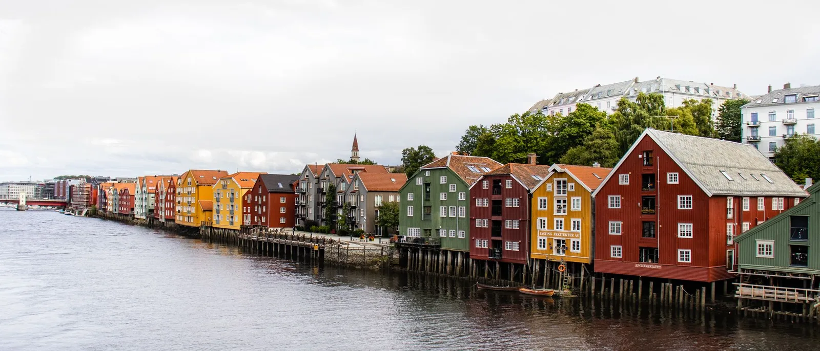Rækken af farvede pakhuse langs Nidelva i Trondheim under en overskyet himmel, floden bred i forgrunden.